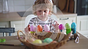 Cute two-year-old boy in bunny ears paint Easter eggs with multi-color paints sitting at the table on the kitchen