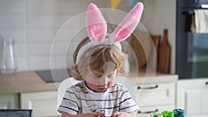 Cute two-year-old boy in bunny ears paint Easter eggs with multi-color paints sitting at the table on the kitchen