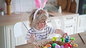 Cute two-year-old boy in bunny ears paint Easter eggs with multi-color paints sitting at the table on the kitchen