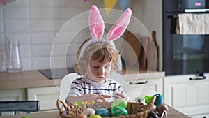 Cute two-year-old boy in bunny ears paint Easter eggs with multi-color paints sitting at the table on the kitchen