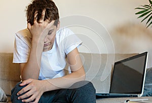 Cute upset boy in white t shirt sitting on the couch in the living room next to laptop and study. Stressed, tired kid.
