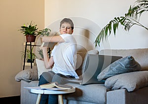 Cute upset boy in white t shirt sitting on the couch in the living room next to laptop and study. Stressed, tired kid