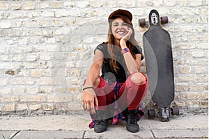 Cute urban girl with longboard sitting sitting next to a brick wall