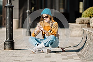 Cute urban girl outdoors with skateboard using smart phone. Happy young woman with skateboard, headphones and cell phone. Hipster