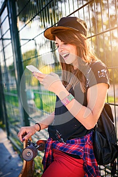 Cute urban girl in skatepark with skateboard using smart phone