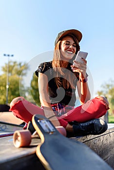 Cute urban girl in skatepark with skateboard using smart phone