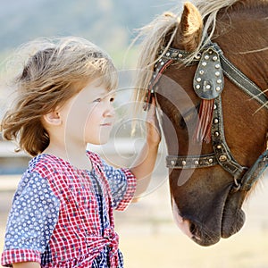 Cute white toddler girl in a rustic style dress caressing brown pony in a field in sunny day