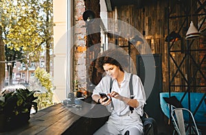 Cute woman with afro hair holding smart phone device, using mobile app sitting at outdoor table in modern city cafe