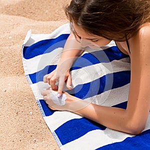 Cute woman is applying sunscreen on her hand with the finger sunbathing on the towel at the beach