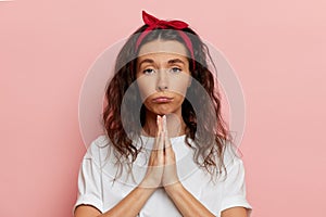 Cute woman begging with cute and upset face, she folded palms together near her face,  over pink background