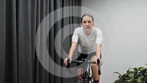 Cute woman cycling indoors on smart trainer wearing white jersey and pink socks. Cardio fitness workouts at home.