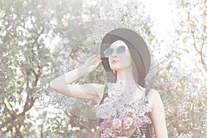 Cute woman in flowered park walks on nature at sunset. Lovely girl in elegant hat in trendy sunglasses in black fashionable dress