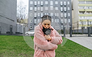 Cute woman in glasses and a pink coat stands on the street with a puppy in her arms, hugging and kissing a pet. Attractive lady