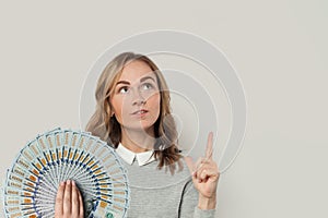 Cute woman in gray sweater pointing up with fan of money dollars against white background