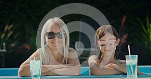 Cute woman with her daughter, relaxing in the pool. Cooling drinks are next to them. Looking at the camera