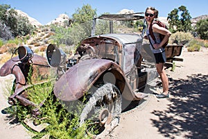 Cute woman hiker poses by an abandoned old fashioned car rusting and decaying in the desert of Joshua Tree National Park