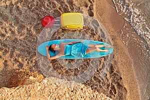 Cute woman lay on surfboard, suitcase and hat on sand. Desire of adventure, summer time, surf and holiday trip idea