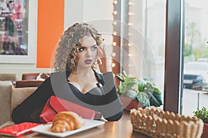 Cute woman with makeup and curly brown hair sitting in cafe and looking at window