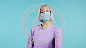 Cute woman with medical mask and violet dyed hairstyle showing thumb up sign over blue background. Positive young girl