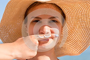 Cute woman is posing with moustache drawn with sun cream on her finger under her nose at the sea background