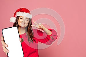 Cute woman in red Santa hat showing smartphone with empty white screen display for new Application on bright pink wall background