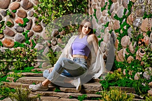 Cute woman sits on stone path surrounded with old overgrown stone wall in the park