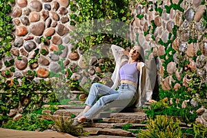 Cute woman sits on stone plate steps with old overgrown stone wall in the park