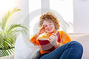 Cute woman sitting on bed in the morning, drinking tea, reading book, casual style, blue jeans, orange sweater, feeling