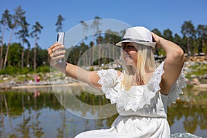 A cute woman takes a selfie and poses on her phone& x27;s camera on the shore of a beautiful forest lake. Lifestyle. In a