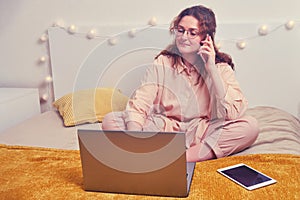 Cute woman in talking on the phone on the home bed and remotely working on a laptop. The girl works at the computer via the