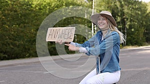 Cute woman try to stop passing car with cardboard poster in forest on roadside of empty road. Lady in hat and denim