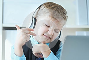 Cute 6 year old boy in suit listening to music or audio tutorial on headphones at the office background.