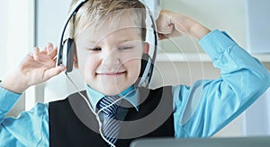 Cute 6 year old boy in suit listening to music or audio tutorial on headphones at the office background.