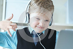 Cute 6 year old boy in suit listening to music or audio tutorial on headphones at the office background.