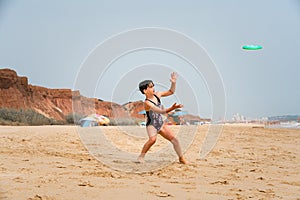 Cute youg girl in swimsuit standing on a beach by the sea throwing a green disc.