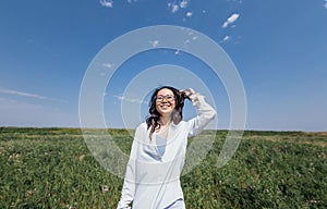 A cute young Asian woman with glasses smiles against a background of blue sky and green dense grass
