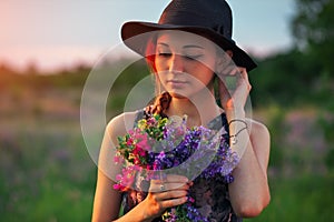 Cute young attractive girl with a bouquet of colorful flowers in her hands and a black hat. Evening walk in nature during sunset.