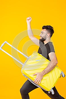 Cute young bearded hipster man holding a heavy yellow suitcase on a yellow background. Concept of heavy load for