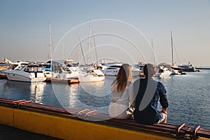 Cute young beautiful couple sitting and hugging together at pier at port, hipsters. Back view