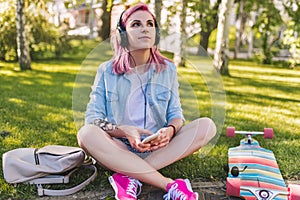 Cute young beautiful woman with pink hair in headphones listening to music dreaming and looking up sitting in park on green grass