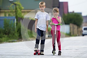 Cute young blond children, brother and sister, girl in pink clothing on scooter and handsome boy on skateboard playing together on