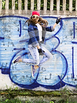 Cute young blonde teenager girl in a baseball cap and jeans shirt jumping against a stone wall background. Hip hop,