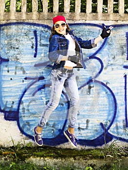 Cute young blonde teenager girl in a baseball cap and jeans shirt jumping against a stone wall background. Hip hop,