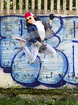 Cute young blonde teenager girl in a baseball cap and jeans shirt jumping against a stone wall background. Hip hop,