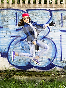 Cute young blonde teenager girl in a baseball cap and jeans shirt jumping against a stone wall background. Hip hop,