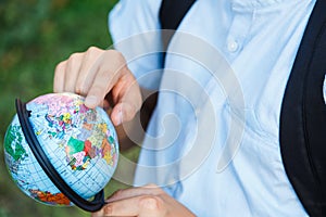 Cute, young boy in blue shirt with backpack and workbooks holds globe in his hands in front of his school. Education