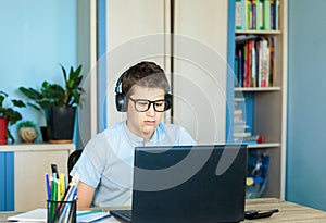 Cute young boy in blue shirt sitting behind desk in his room next to laptop and study. Teenager in earphones makes homework,