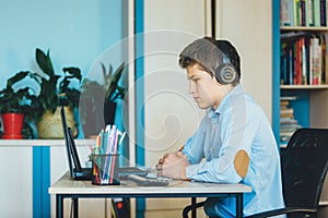 Cute young boy in blue shirt sitting behind desk in his room next to laptop and study. Teenager in earphones makes homework,