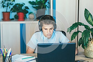 Cute young boy in blue shirt sitting behind desk in his room next to laptop and study. Teenager in earphones makes homework,