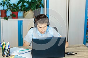 Cute young boy in blue shirt sitting behind desk in his room next to laptop and study. Teenager in earphones makes homework,